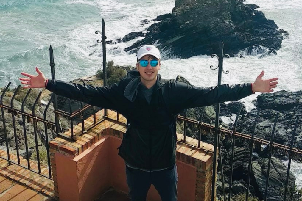 Brett Stievater on a cliffside in Spain wearing sunglasses and a baseball cap with his arms opened wide smiling