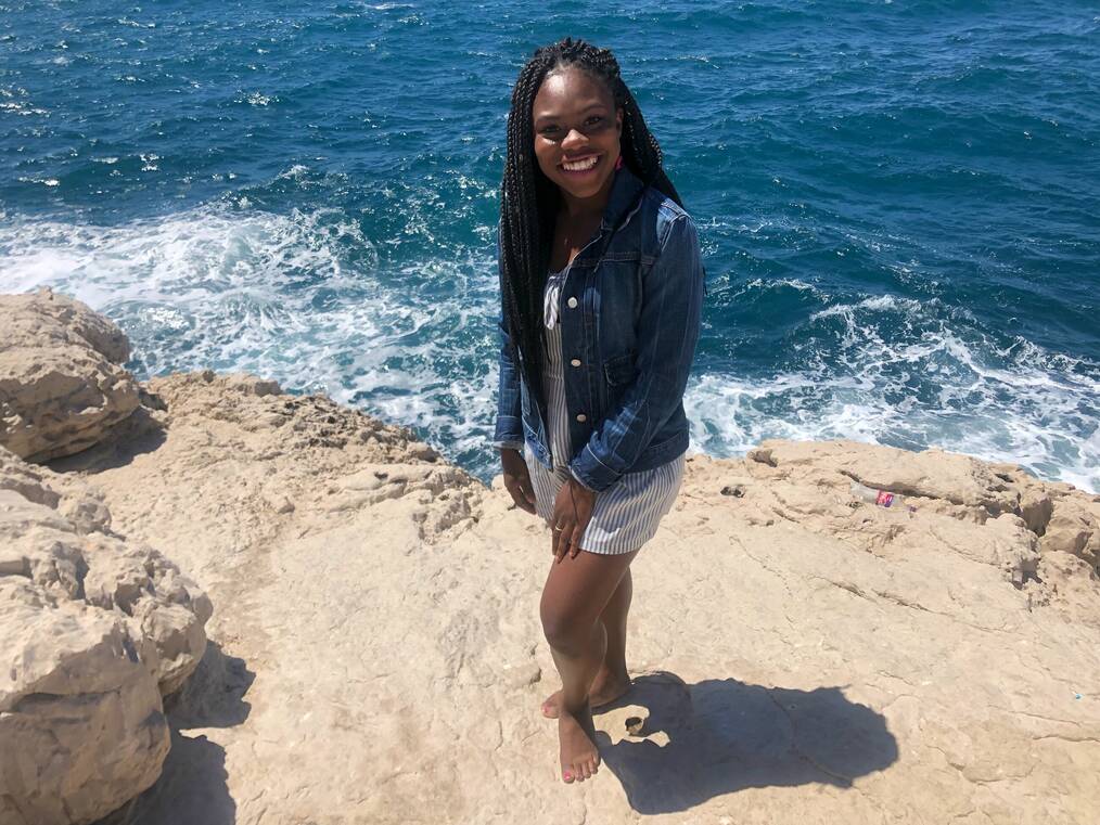 A woman poses smiling in front of an ocean.