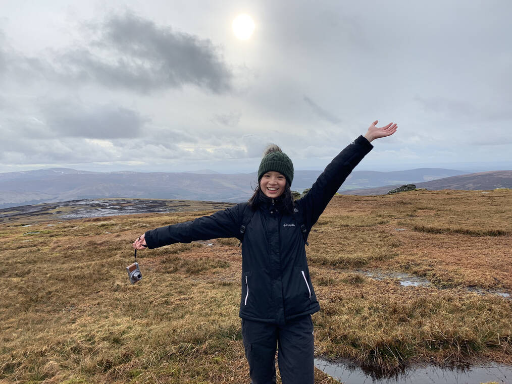 A woman poses in a field with her arms in the air.