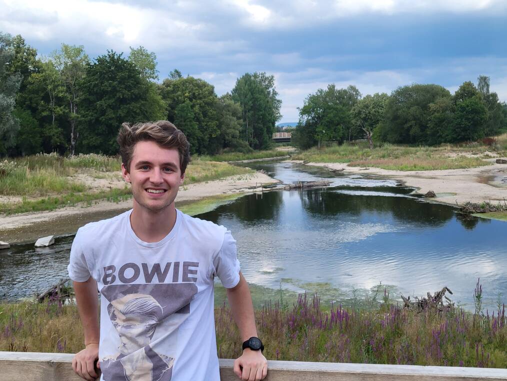 Ian A. standing in front of a lake in Germany smiling wearing a white Bowie shirt 