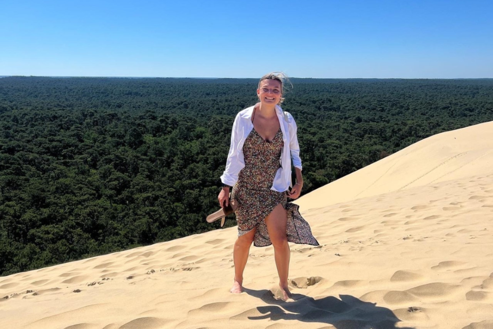 Jessica Bertolini standing in the sand on the beach wearing a dress and a white cardigan with blonde hair