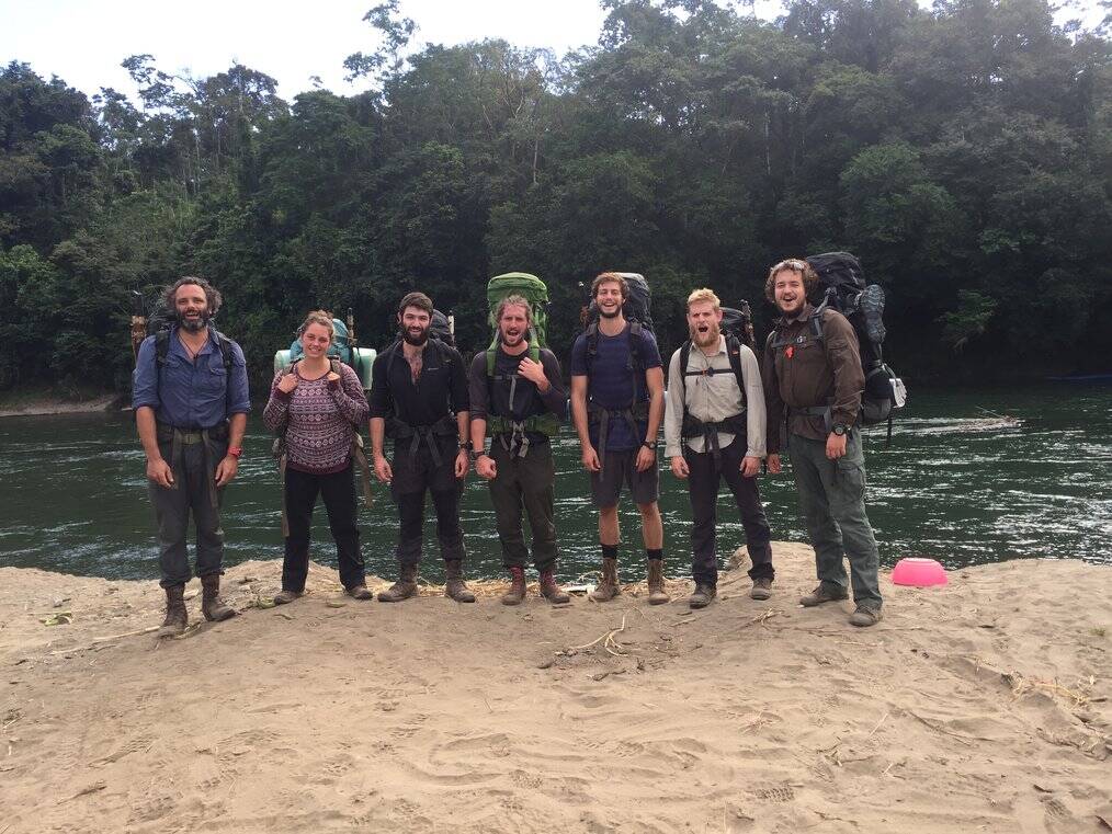 A group of people stand on a riverbank with large backpacks.