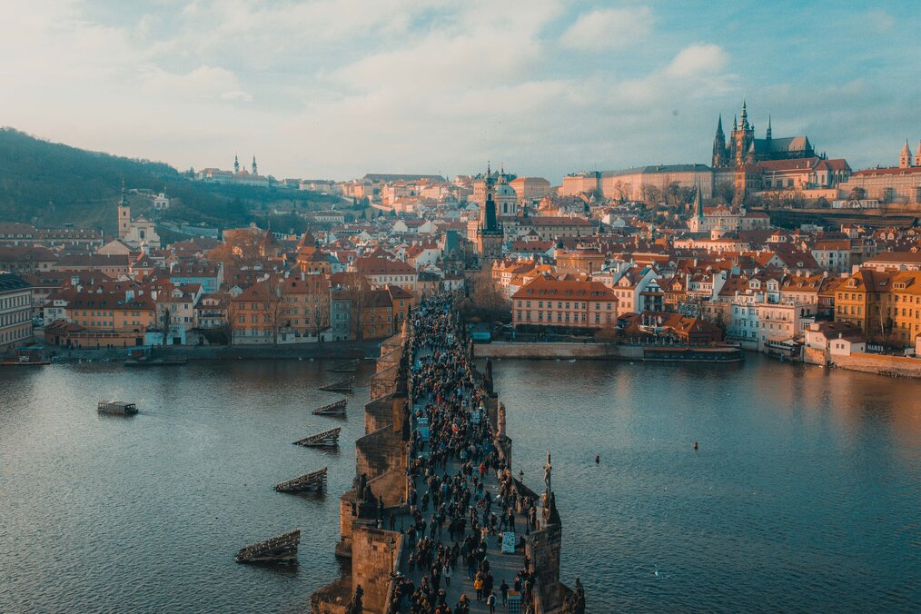 Prague A drone shot of the city of Prague shows a bridge over a river leading to buildings.