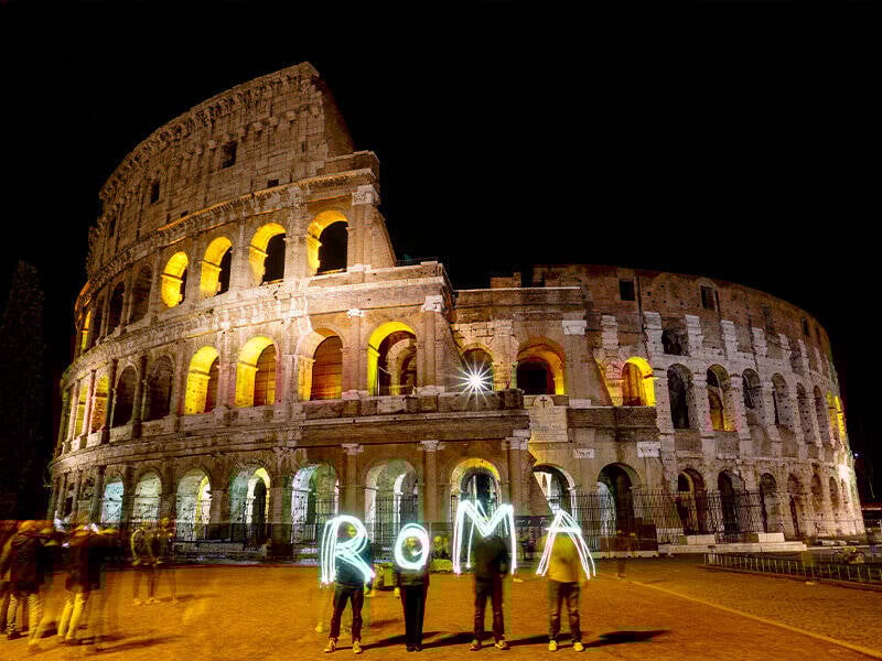 The neon word "ROMA" glows in the air in front of the Colosseum in Rome.