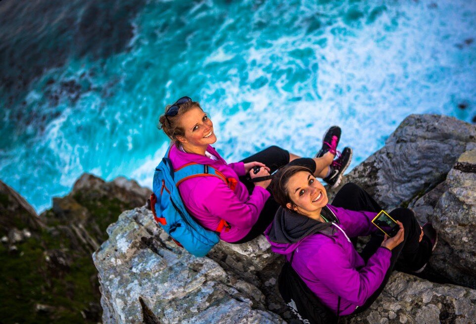VACorps Two women sitting on a cliff by the ocean look up at and smile for the camera.