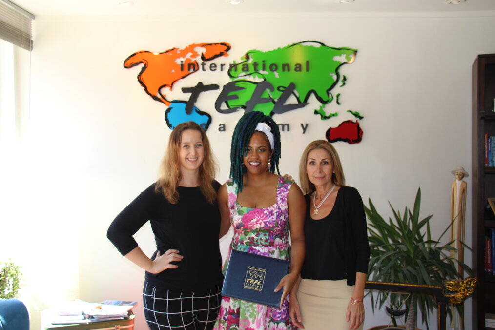 Three women stand together smiling. The one in the middle holds a diploma.