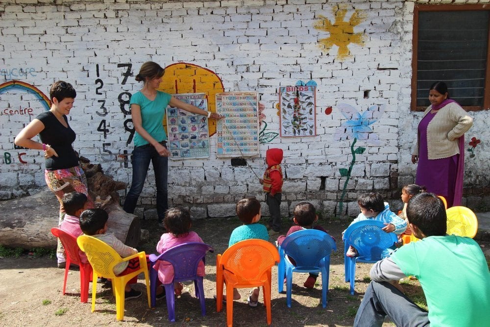 PMGY Children sit around in chairs while a teacher points at a poster on the wall.