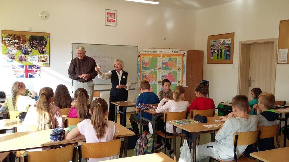 GV Two older adults stand at the front of a classroom of children.