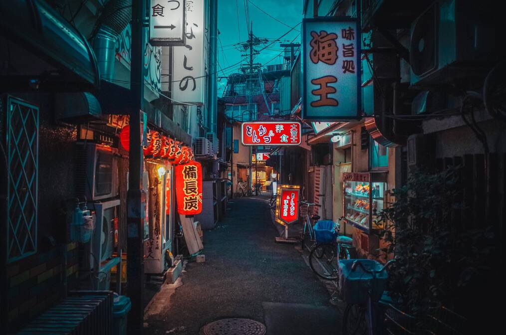A darkened alleyway is lit up by neon signs in Japanese.