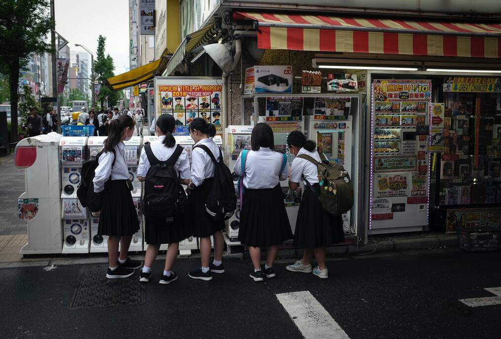 Girls in school uniforms stand in front of a store kiosk with their backs to the camera.