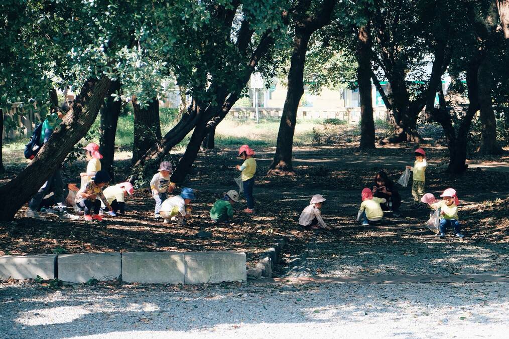 Japanese children wearing hats crouch in the leaves under some trees.