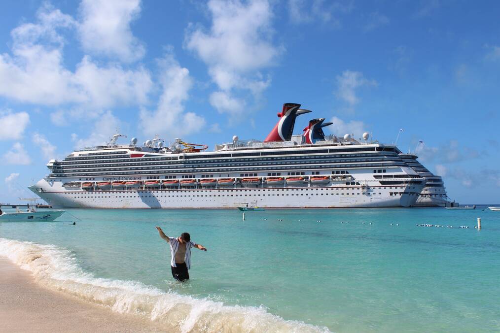 A man stands in the water on the beach with a cruise ship in the background.