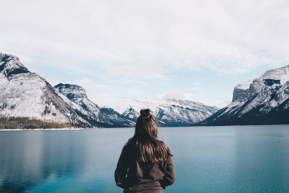 A woman stands with her back to the camera looking out to a lake.