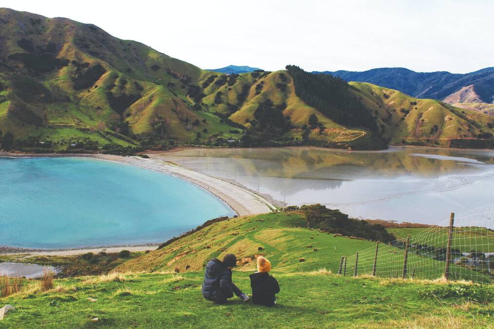 Two people sit on a hill overlooking a lake and mountains.