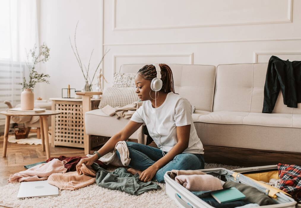 A young woman packs her things in her suitcase while listening to music in her living room