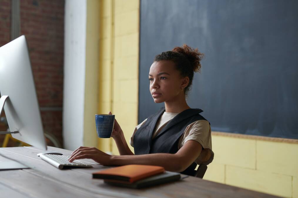 A woman holding a mug sits and looks at a computer monitor.