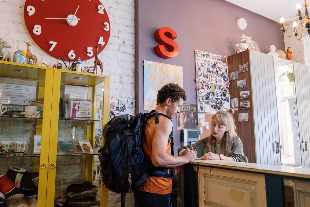 A woman at a hostel front desk helps a man wearing a backpack.