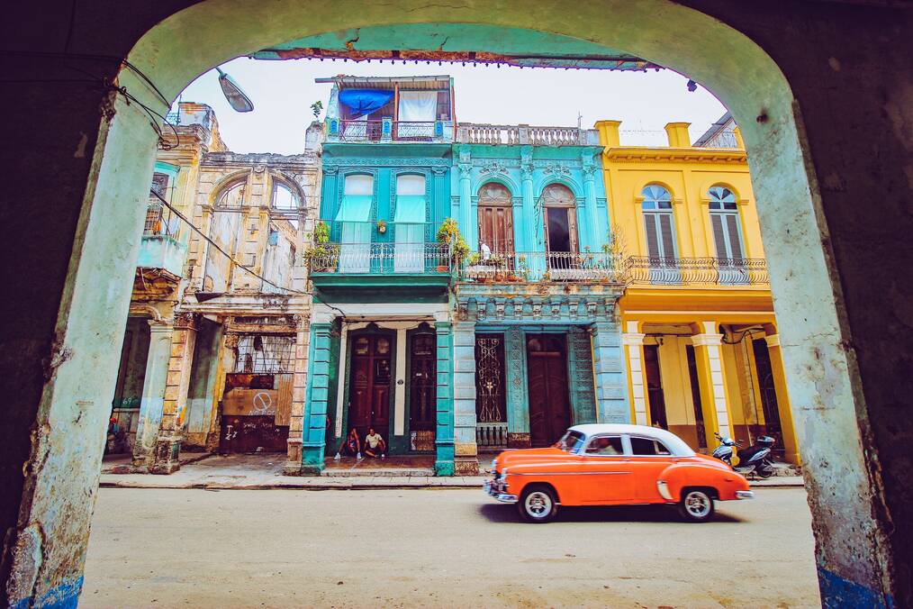 An orange classic car drives by colored buildings in Havana, Cuba.