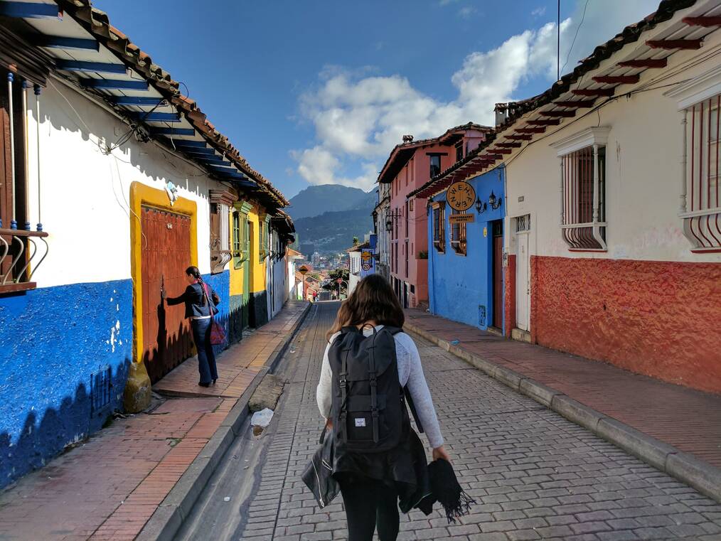 A woman wearing a backpack walks down a street with colorful buildings.