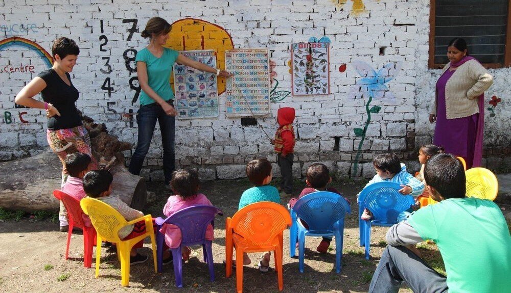 PMGY Children sit around in chairs while a teacher points at a poster on the wall.