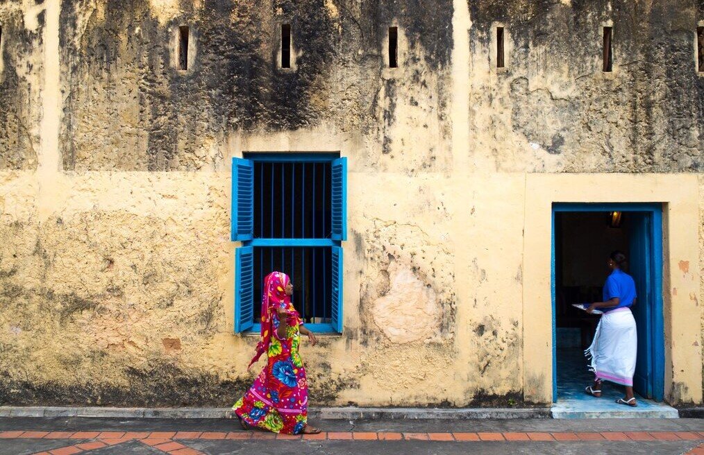 Photo by Giacomo Berardi on Unsplash A woman in a bright pink outfit walks by a window trimmed in bright blue.