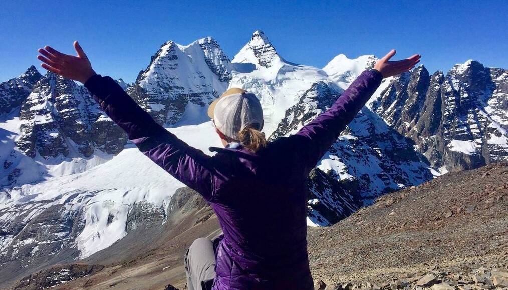 A woman sits with her back to the camera and arms raised in front of a snowcapped mountains.