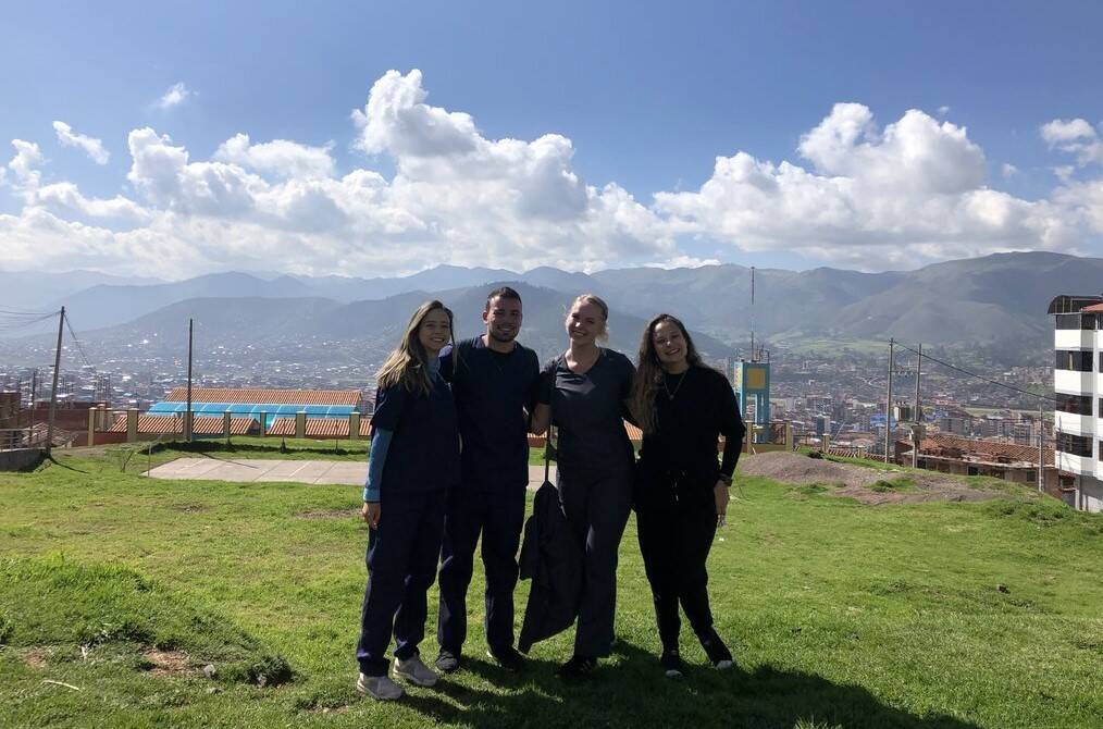 Four people pose for a photo standing on green grass with mountains in the background.