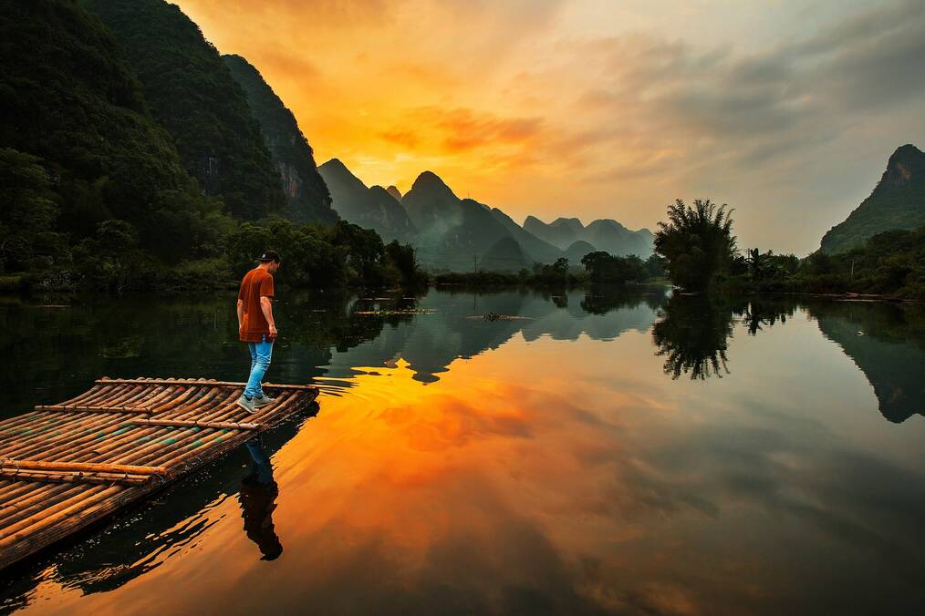 A man stands at the end of a dock with mountains in the background.