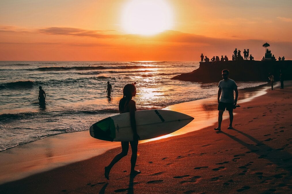 A woman carrying a surf board walks on a beach at sunset.