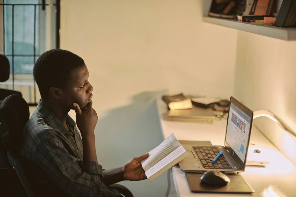 A man reads a book near a laptop.