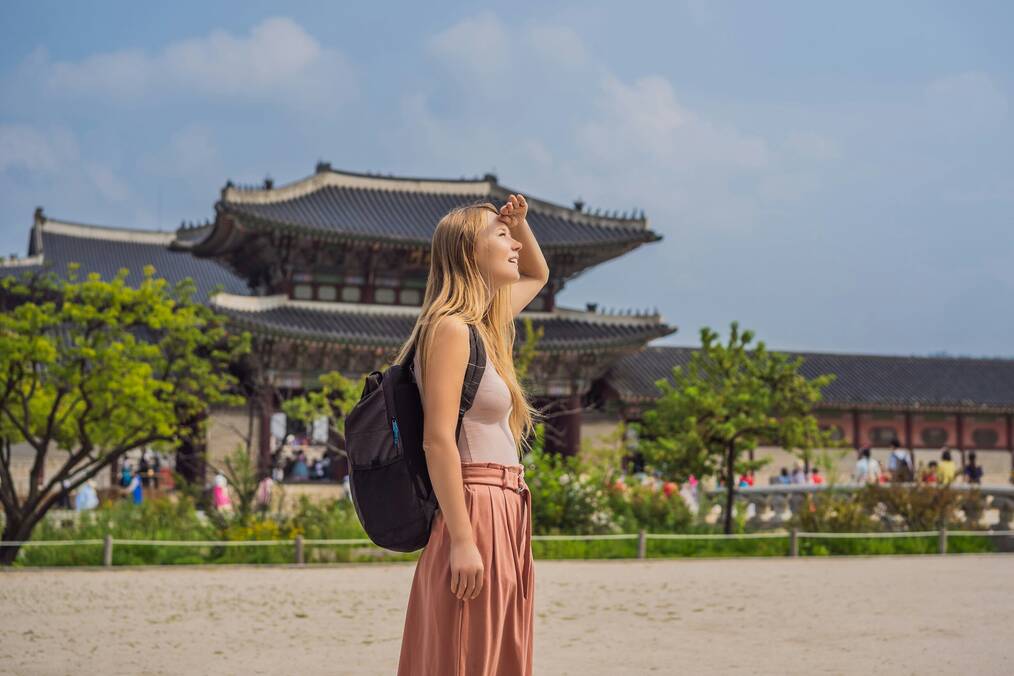 A woman stands in front of a Korean temple as she shields her eyes from the sun.