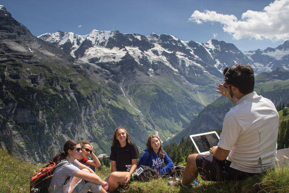 A group of teens outside with mountains in the background look at a teacher holding a tablet.