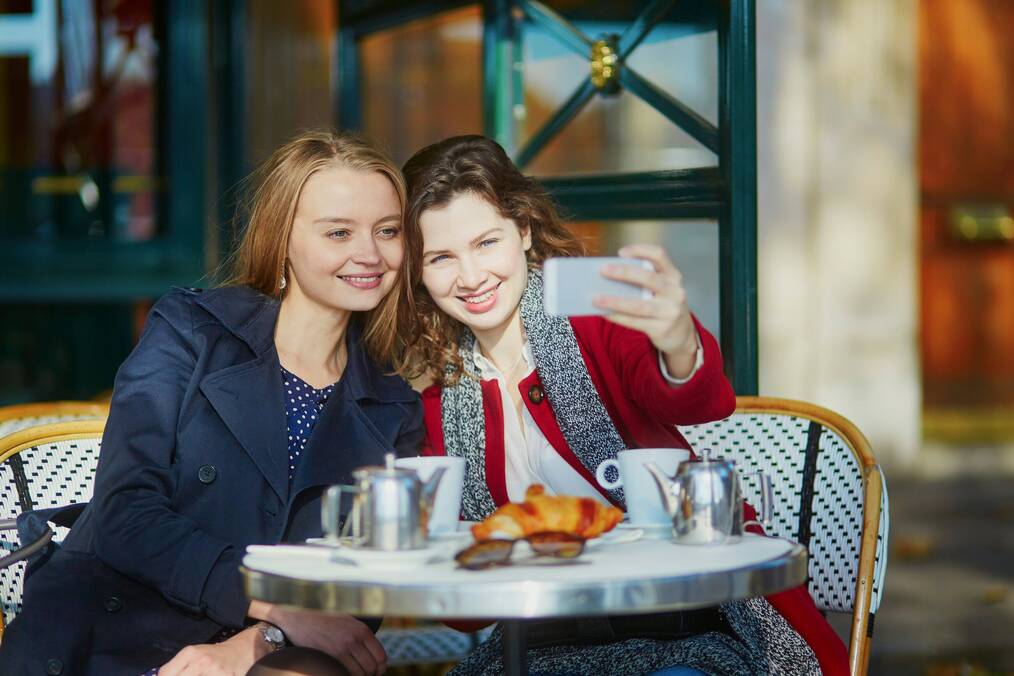 Two women take a selfie at a cafe table with a croissant on the table.
