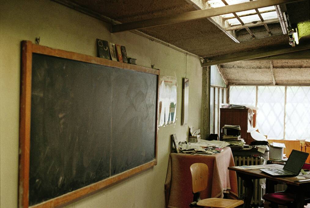 A laptop sits on a desk with a blackboard in the background.
