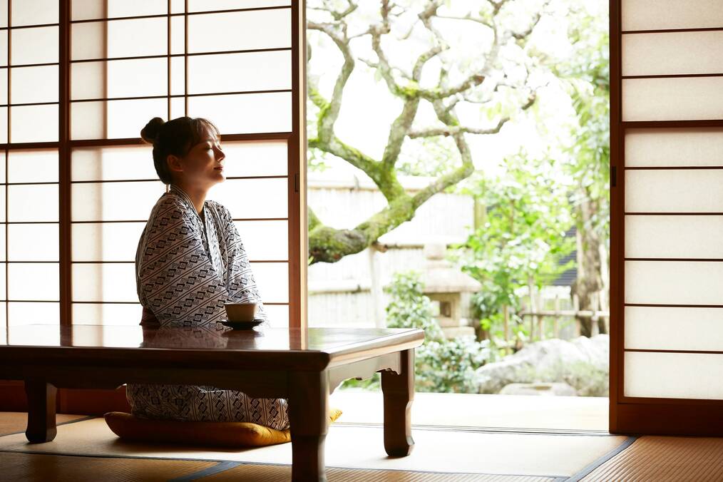 A woman in a kimoni sits on a mat near an open door.