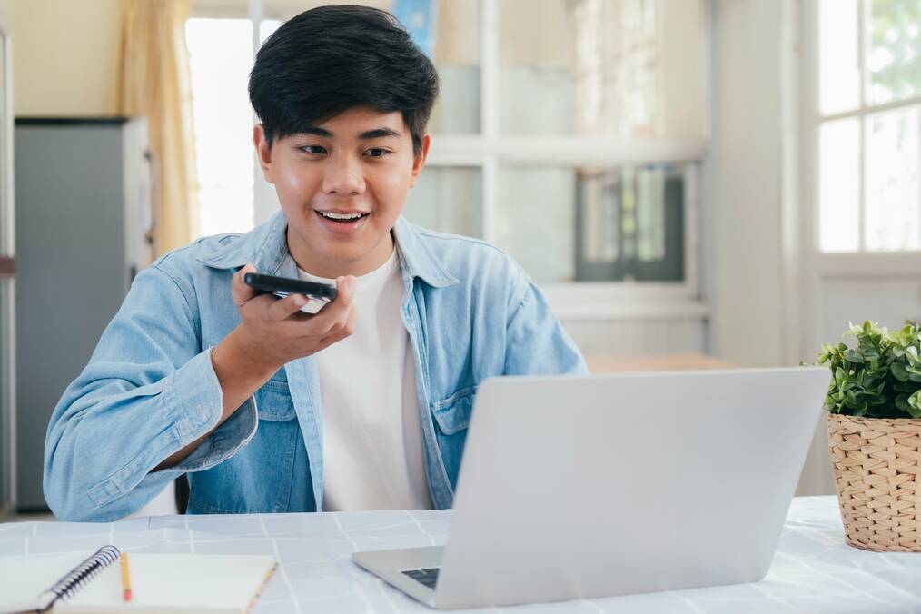 A man records his voice on a cell phone while sitting in front of a laptop.