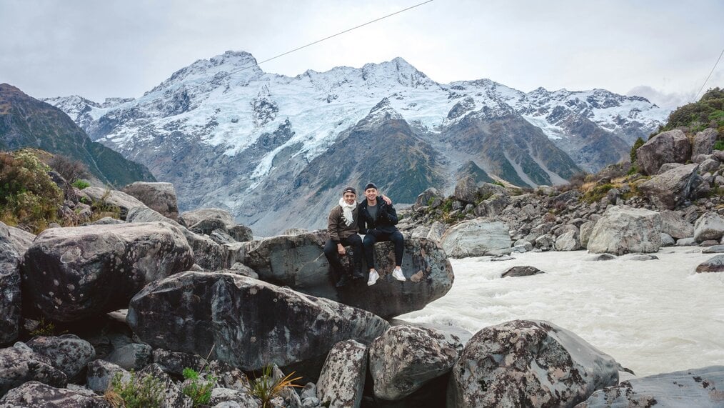 Two men sit on a rock with mountains in the background.