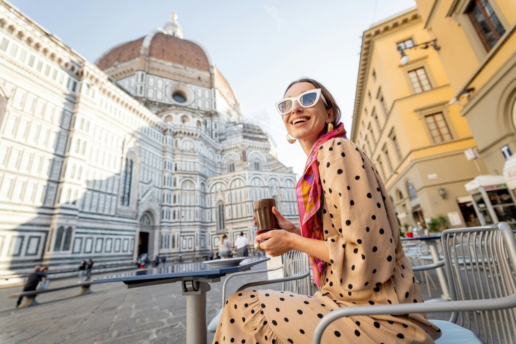 Unsplash woman A woman sits with a coffee outside in Italy with large buildings in the background.