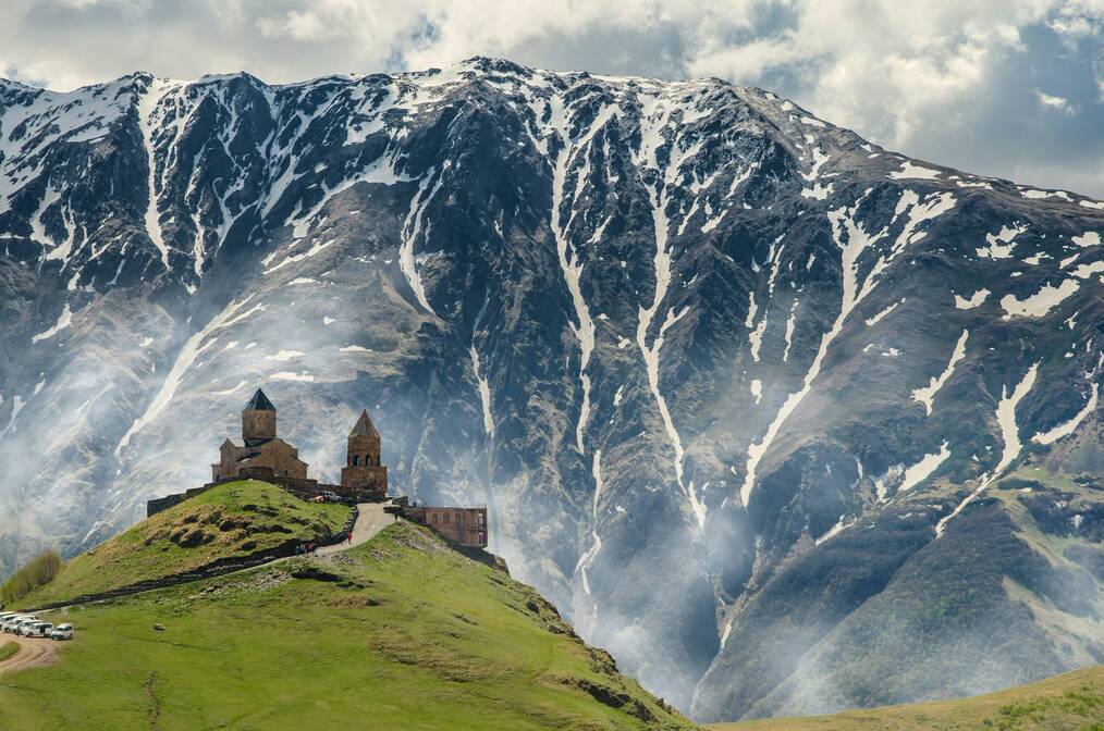 A castle sits on a hill in front of mountains in Georgia.