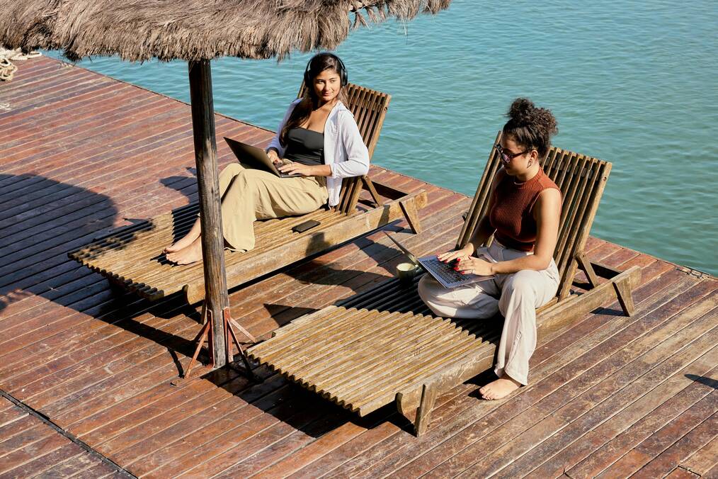 Two women sit with their laptops by the water.
