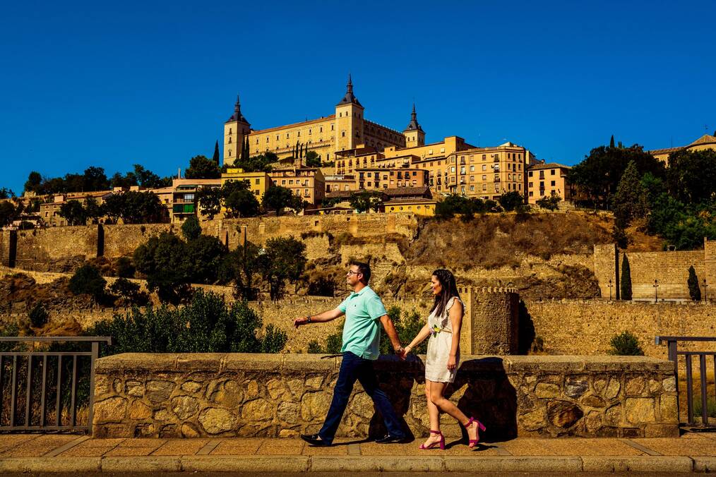 A man and woman hold hands and walk down the street with a castle in the background.