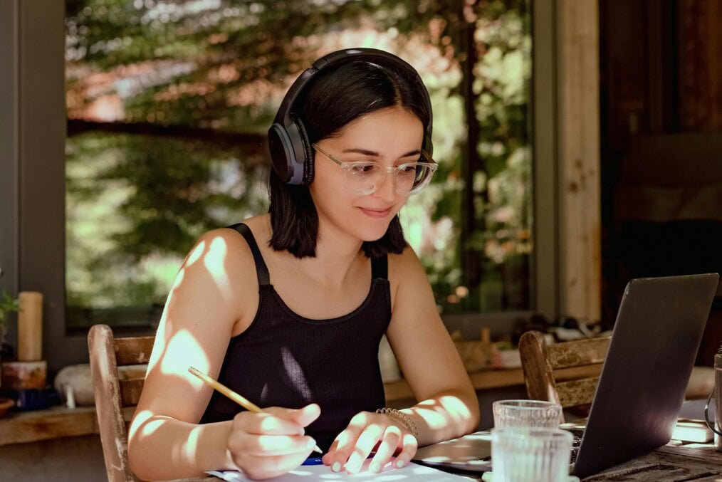 A woman holding a pencil and wearing headphones smiles at a computer screen.