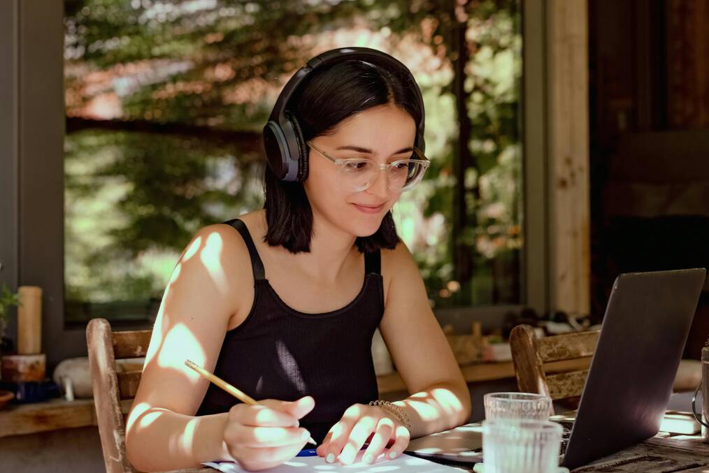 A woman holding a pencil and wearing headphones smiles at a computer screen.