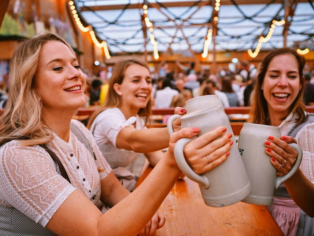 Three women in traditional dress cheers their mugs at Oktoberfest.