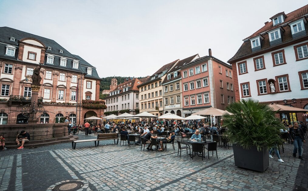 Photo by Ruben Hanssen on Unsplash People sit outside at tables with umbrellas in a German square.