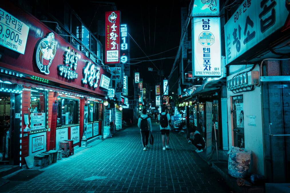 Two people walk down a street in Seoul illuminated by neon signs.