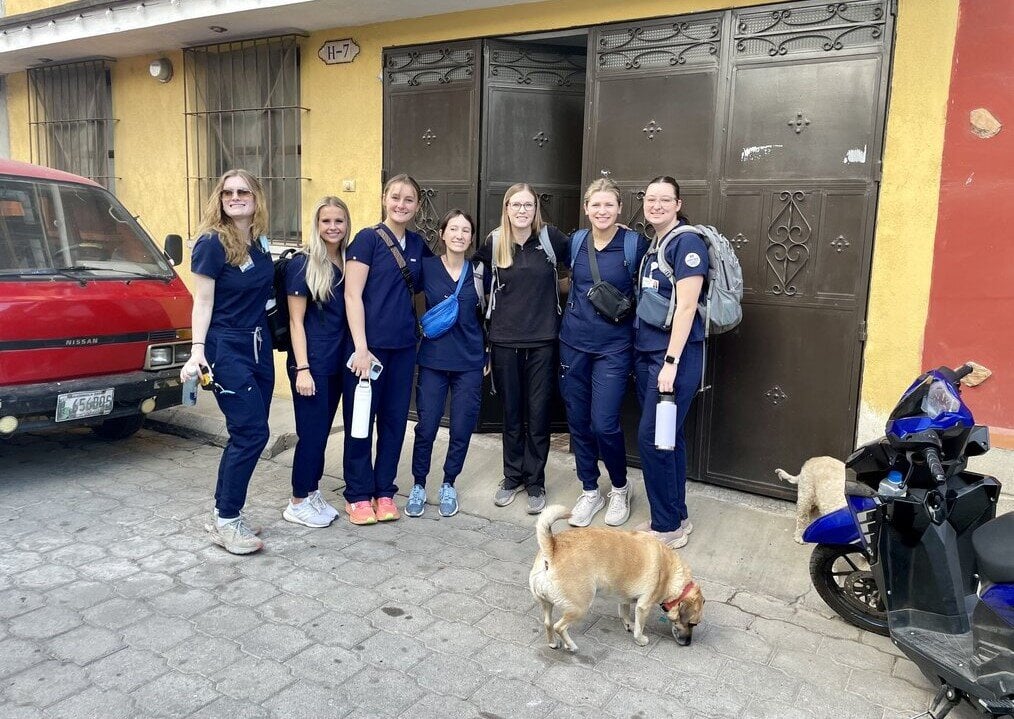 MN A group of women in dark blue scrubs stand outside of a building.