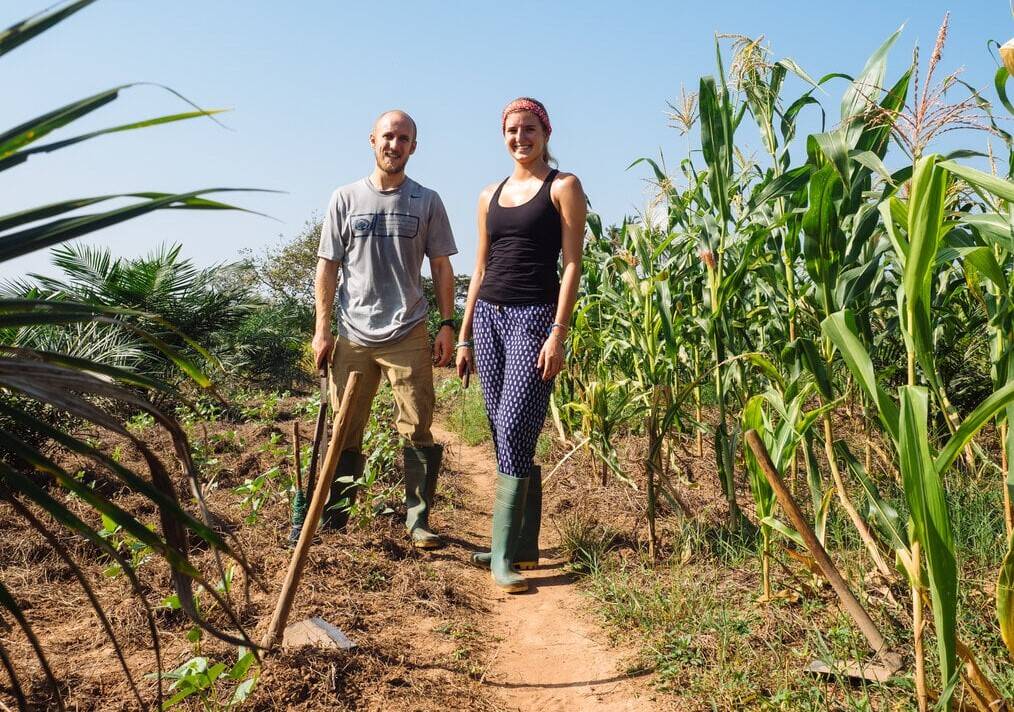 Two volunteers stand and smile next to corn stalks and tools.