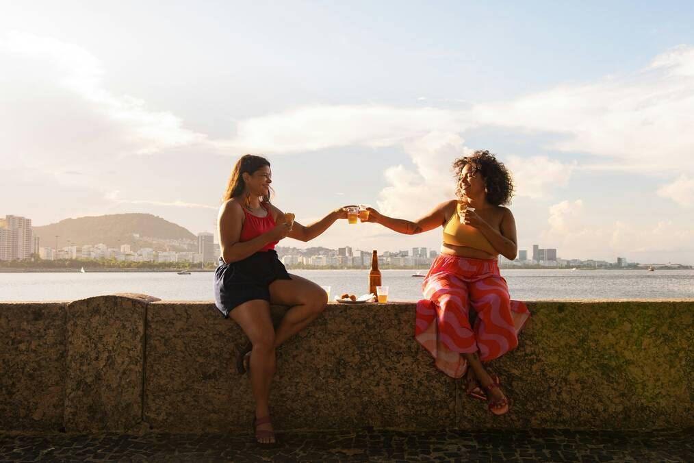 Two smiling women sit on a wall and toast their glasses.
