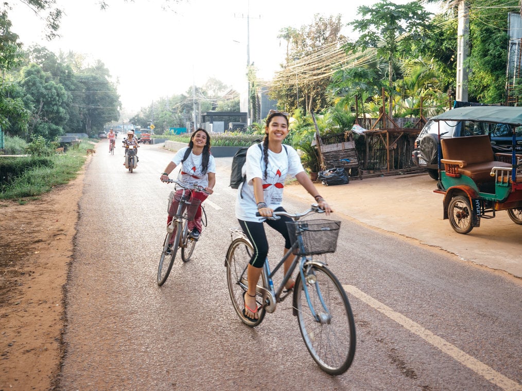 Two young women ride bikes down a road in Cambodia.